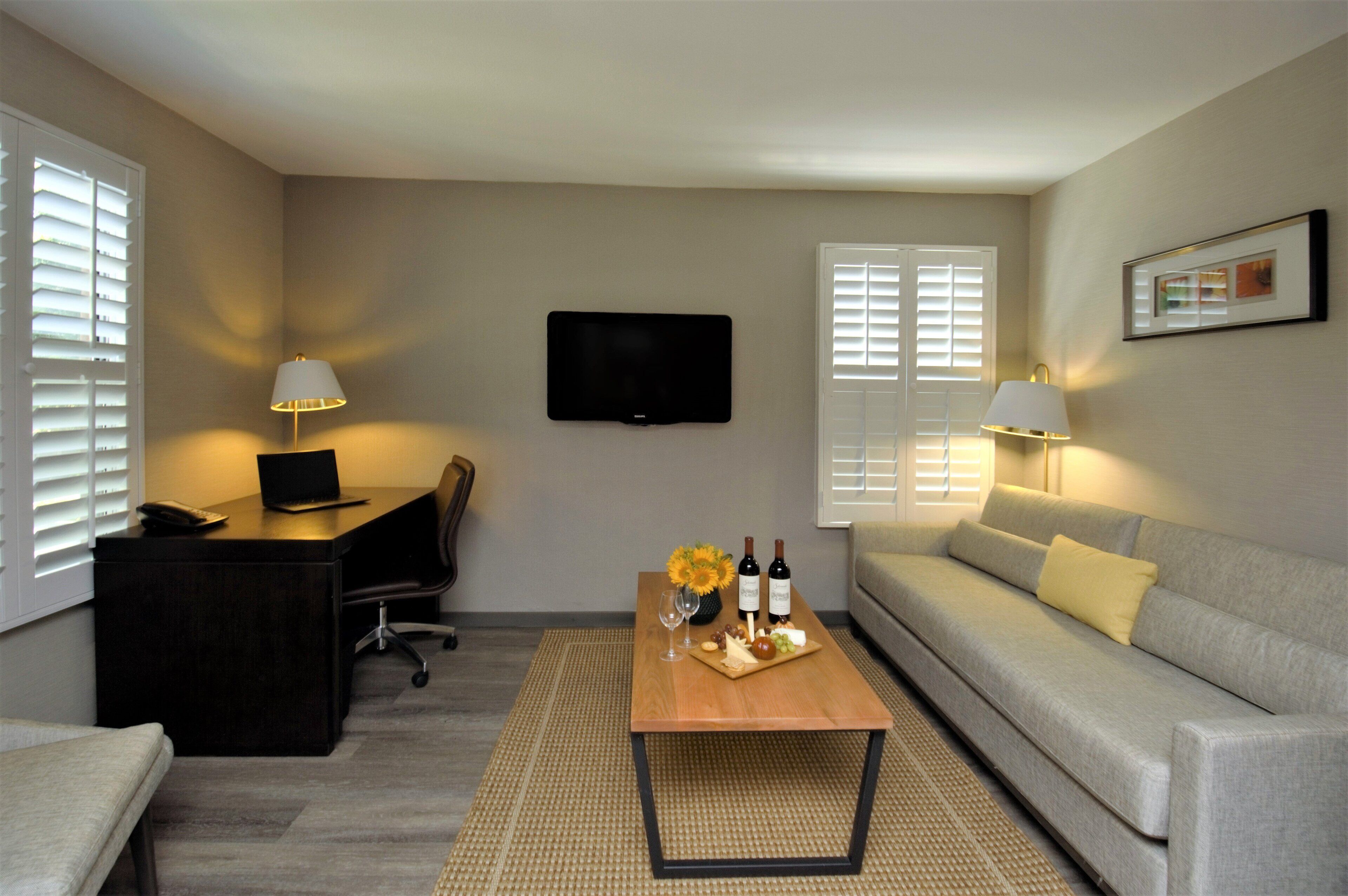 A modern living room with a flat-screen TV mounted on the wall, a desk with a laptop, a sofa, and a coffee table with a tray of snacks and drinks.