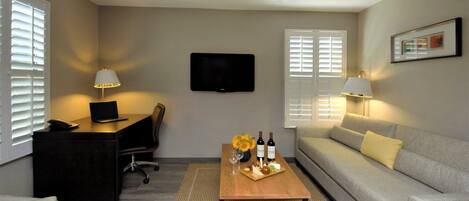 A modern living room with a flat-screen TV mounted on the wall, a desk with a laptop, a sofa, and a coffee table with a tray of snacks and drinks.