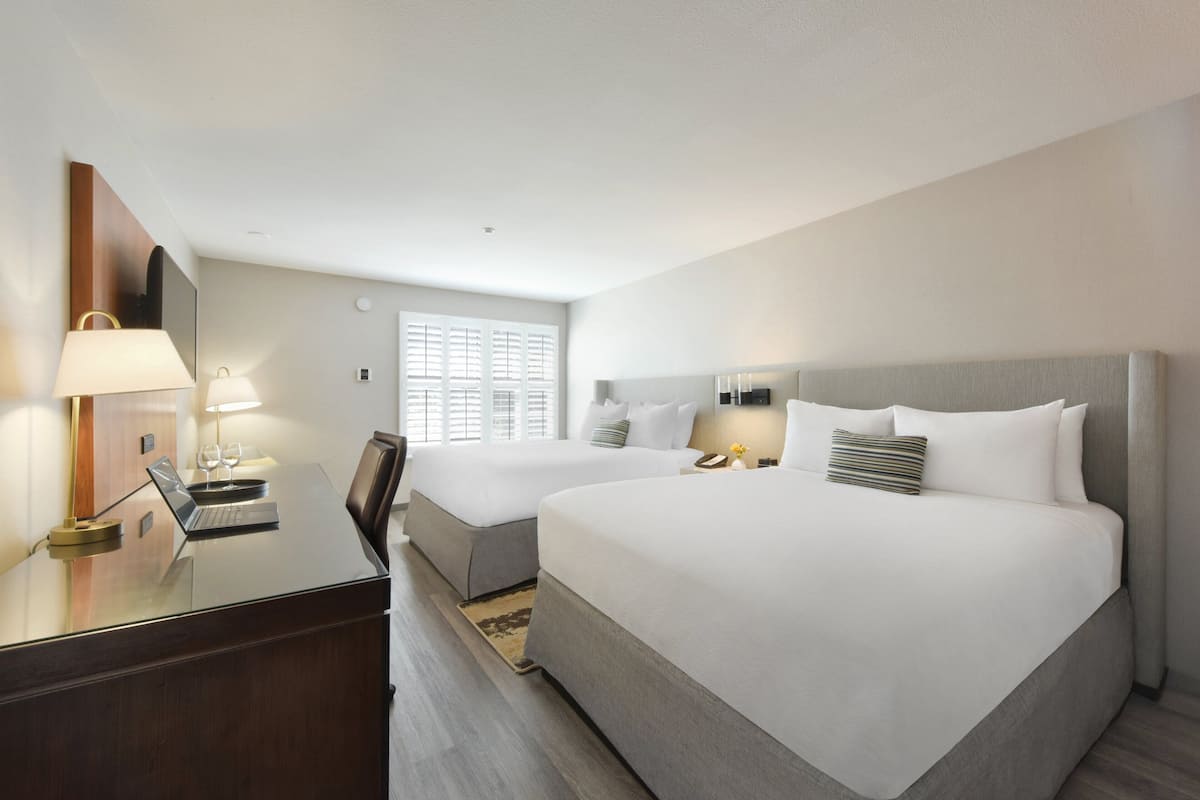 A hotel room with two beds, a desk with a laptop, and a window with blinds.