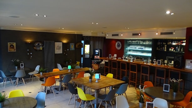 A modern bar area with multiple seating options including tables and chairs, a bar counter with a variety of bottles and glasses, and a television screen. - Sleep & Go Brussels Expo (Grimbergen)