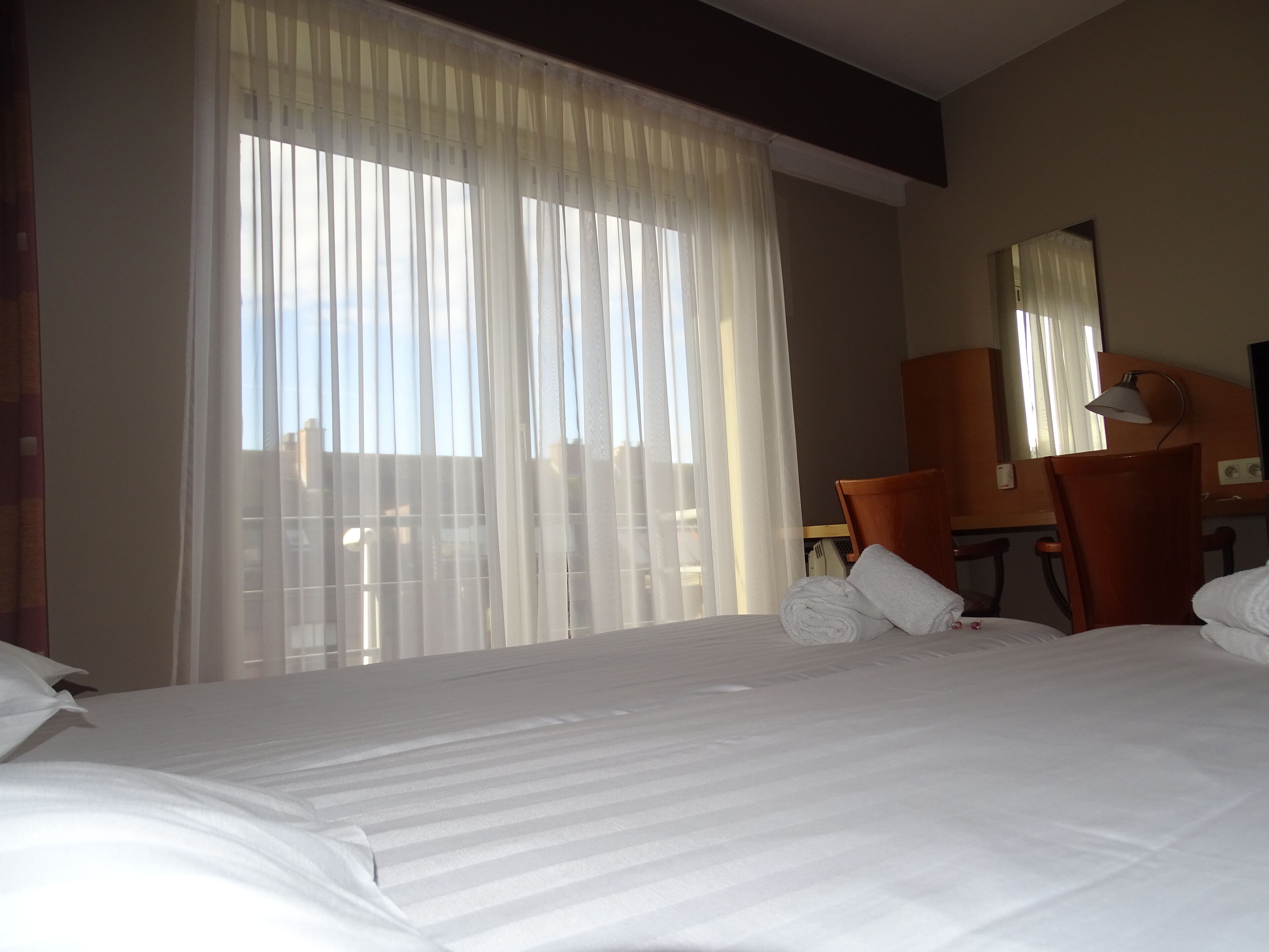 A hotel room with a large bed, a desk, a chair, and a window with sheer curtains.