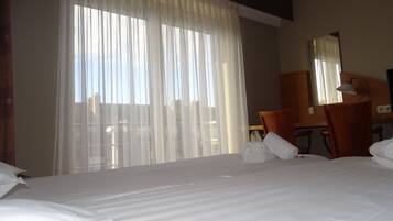 A hotel room with a large bed, a desk, a chair, and a window with sheer curtains.