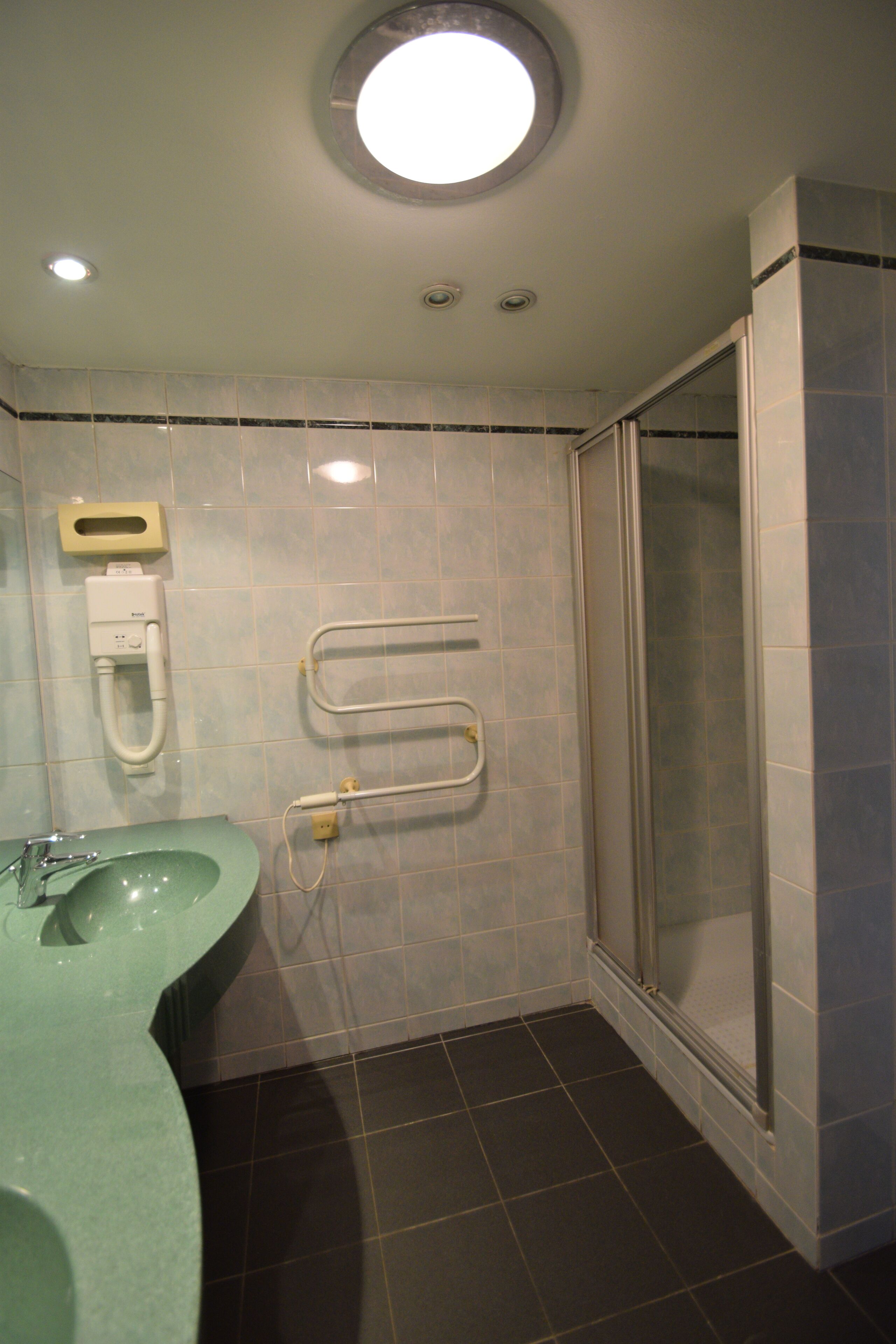 Une salle de bain avec un lavabo vert, un distributeur de papier toilette, un sèche-mains et une cabine de douche en verre.