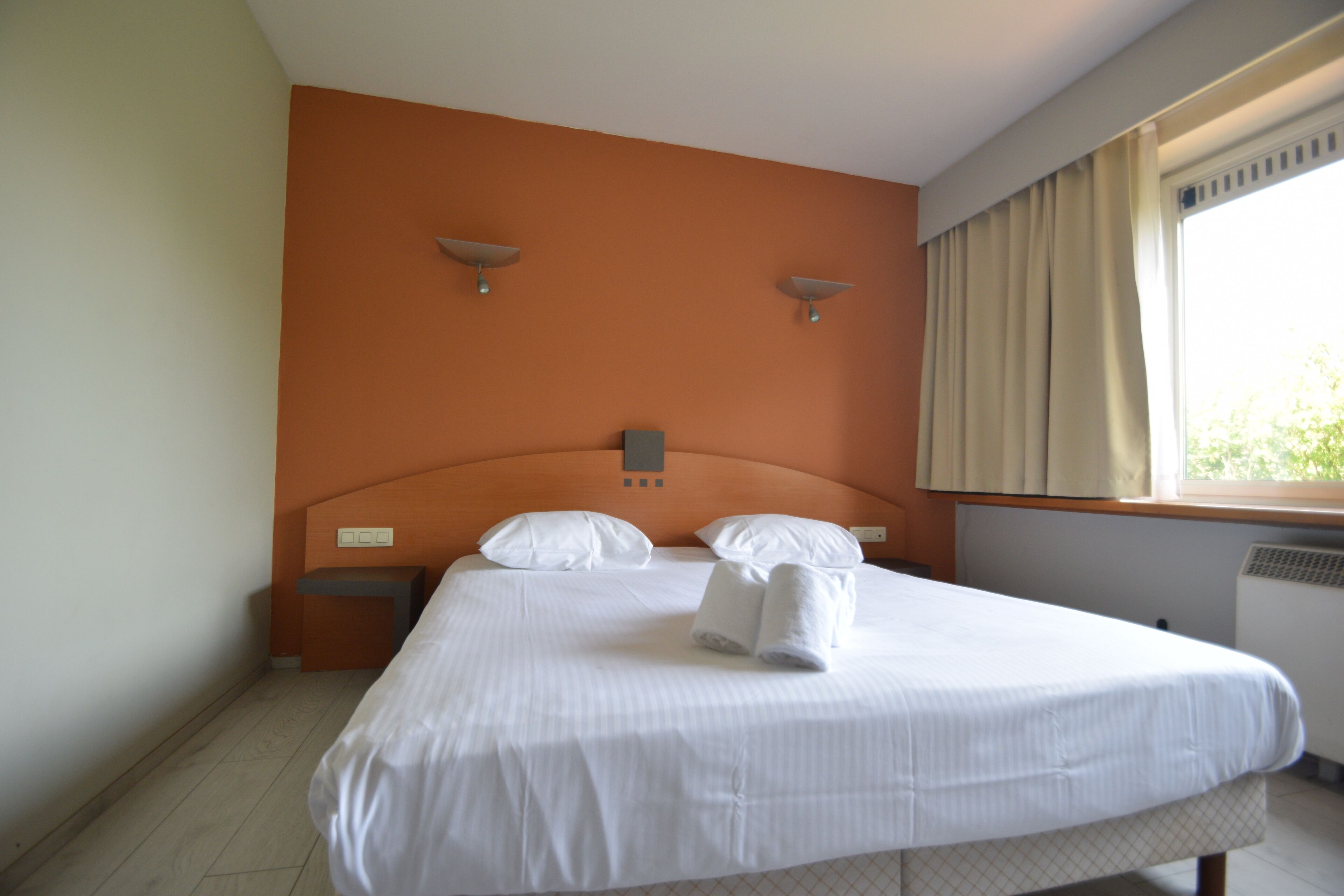 Una cama bien tendida con sábanas blancas y dos almohadas, una mesita de noche con una lámpara, y una ventana con cortinas.