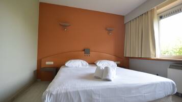 Una cama bien tendida con sábanas blancas y dos almohadas, una mesita de noche con una lámpara, y una ventana con cortinas.