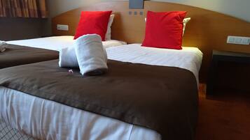 A hotel room with two beds, each with a brown and white bedspread, red pillows, and a white towel on one bed. There is a wooden headboard, a small nightstand with a lamp, and a wall-mounted light fixture.