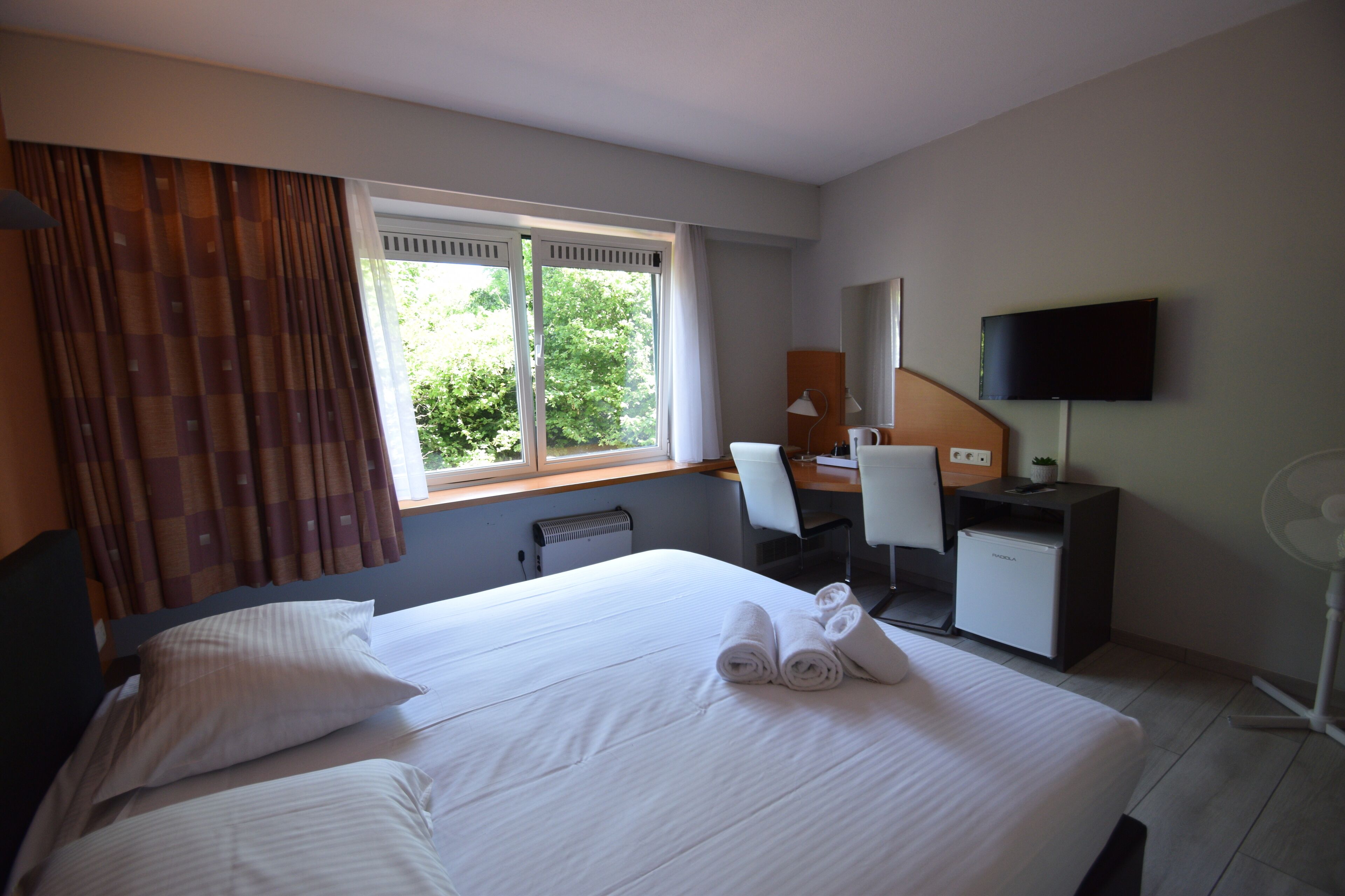 Una habitación de hotel con cama, escritorio, silla, televisor y ventana con cortinas.
