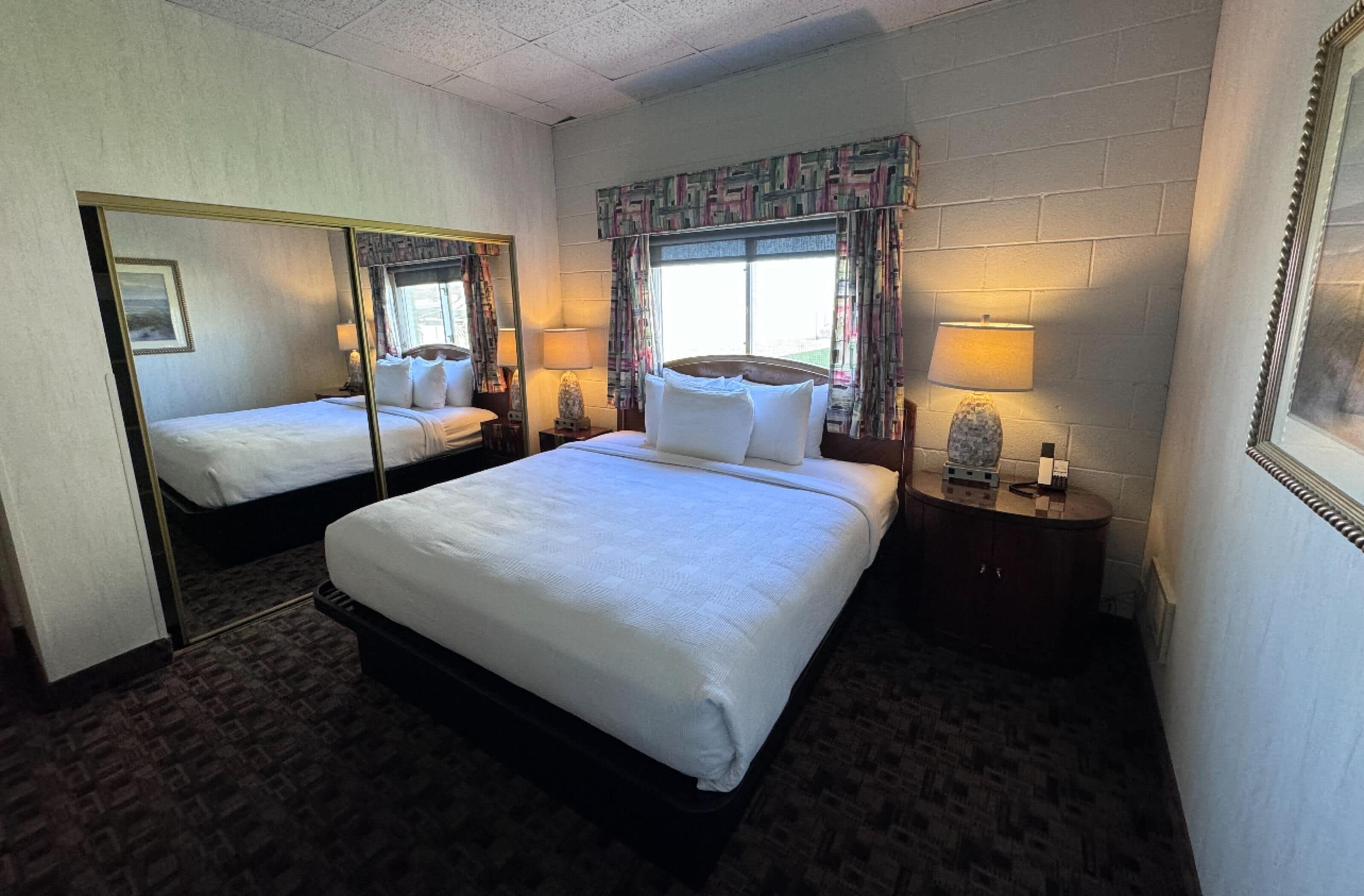 A hotel room with two beds, a large mirror, a window with curtains, bedside lamps, and a small table with a lamp.