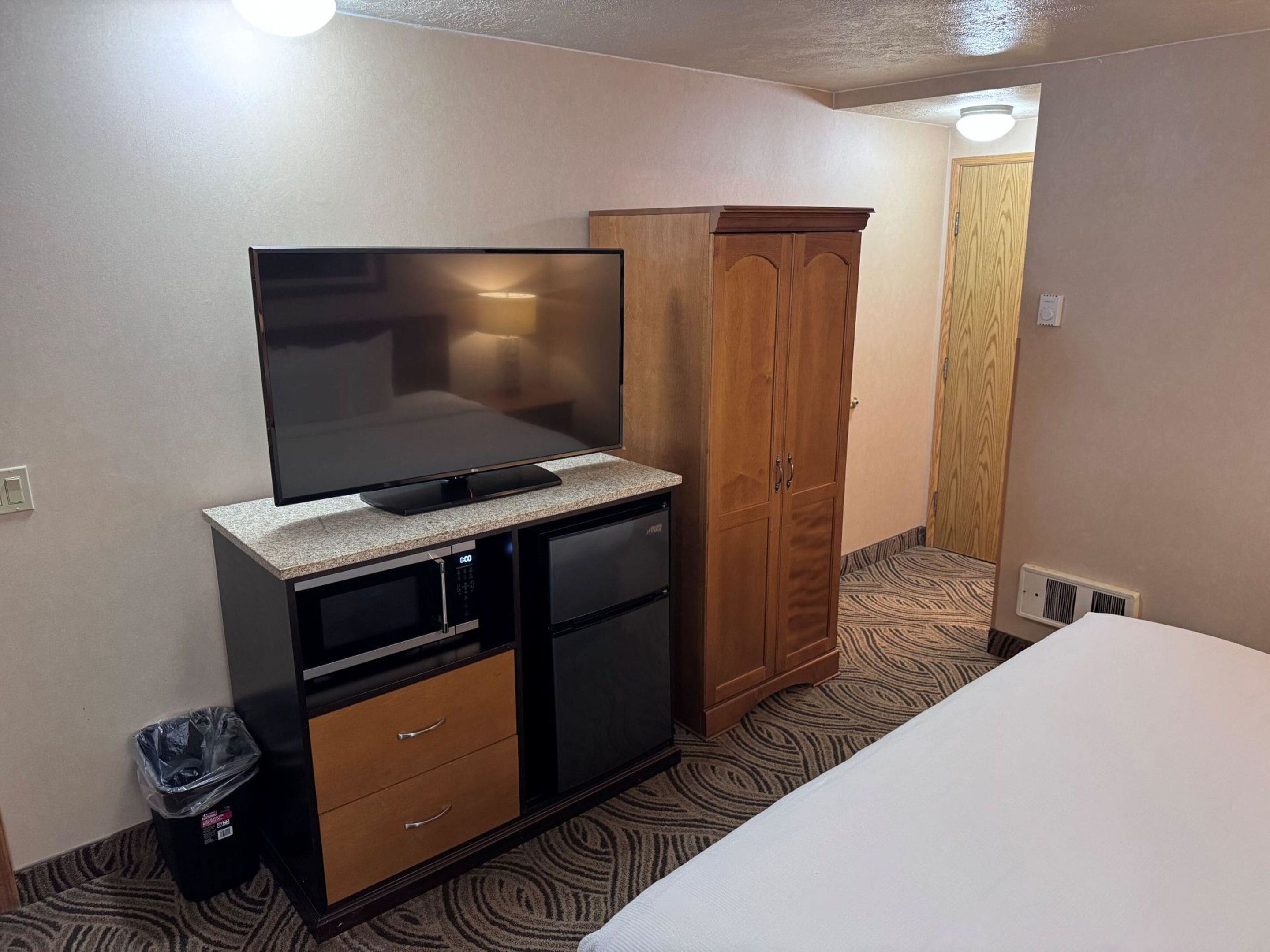 Habitación de hotel con televisión de pantalla plana en un mueble, armario de madera, cesto de basura y cama.