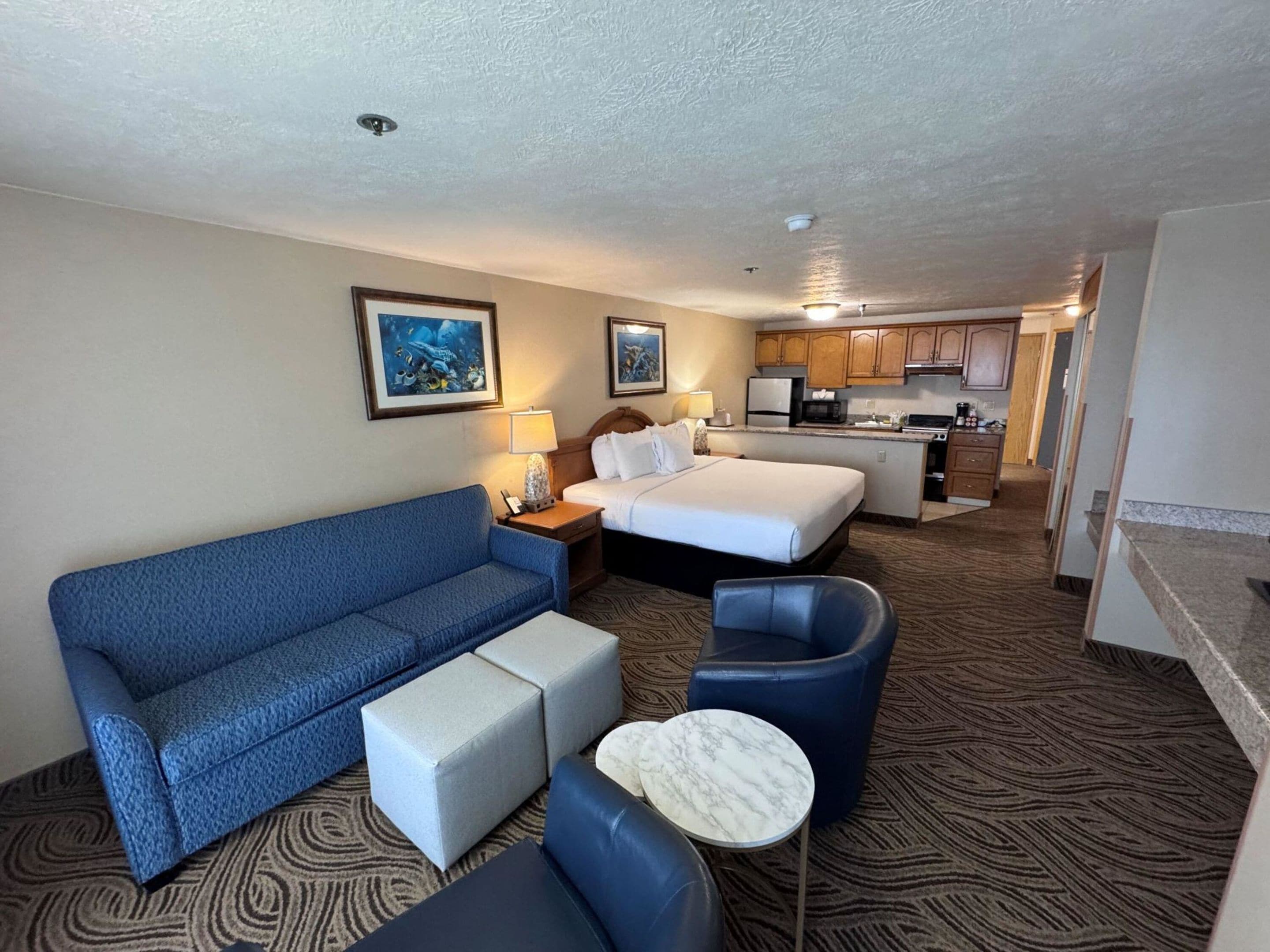 Ein Hotelzimmer mit blauer Couch, einem Bett, einem kleinen runden Tisch und einer Küchenzeile im Hintergrund.
