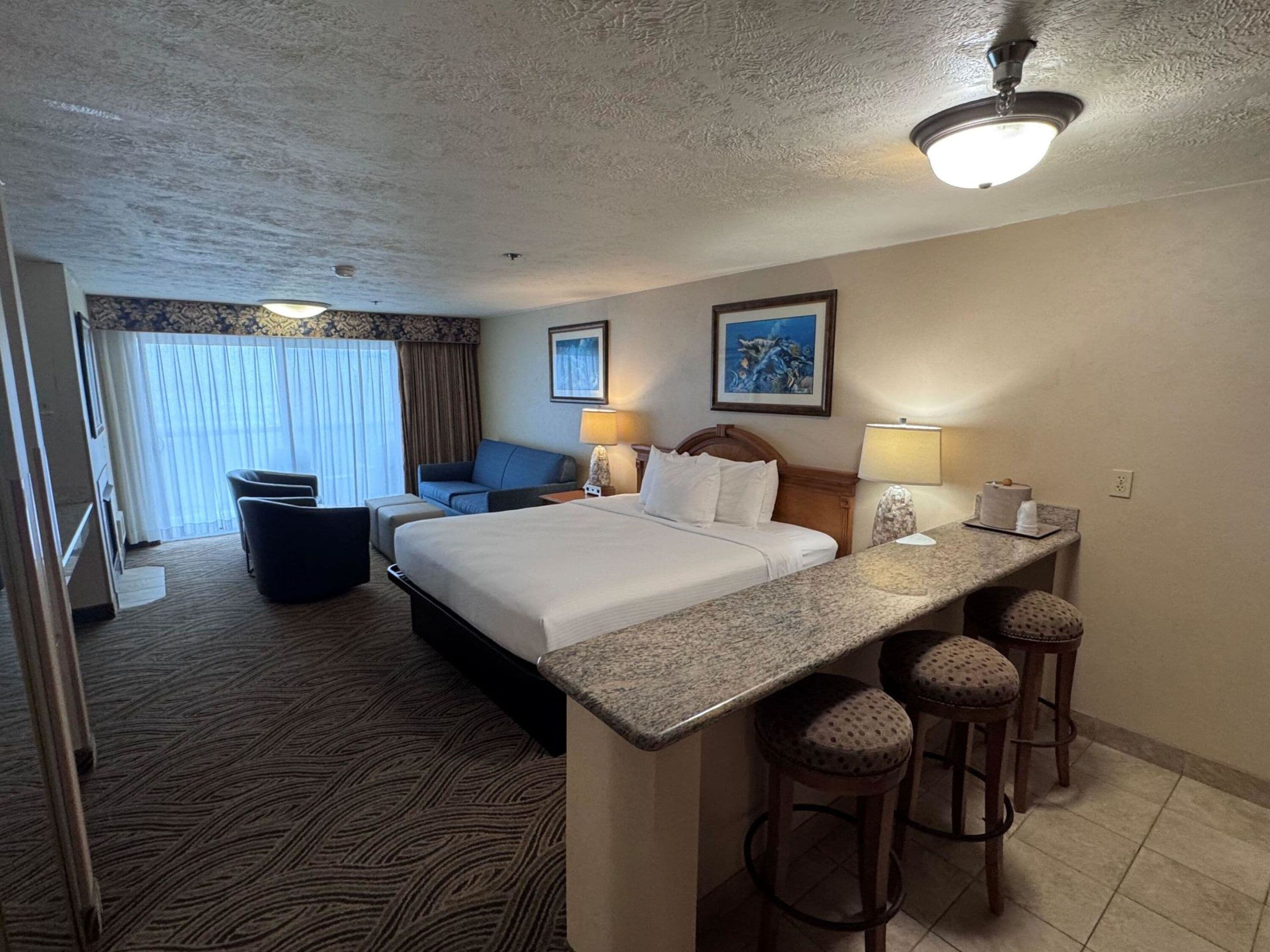 A hotel room with a bed, a desk with stools, a chair, a sofa, and a window with curtains.