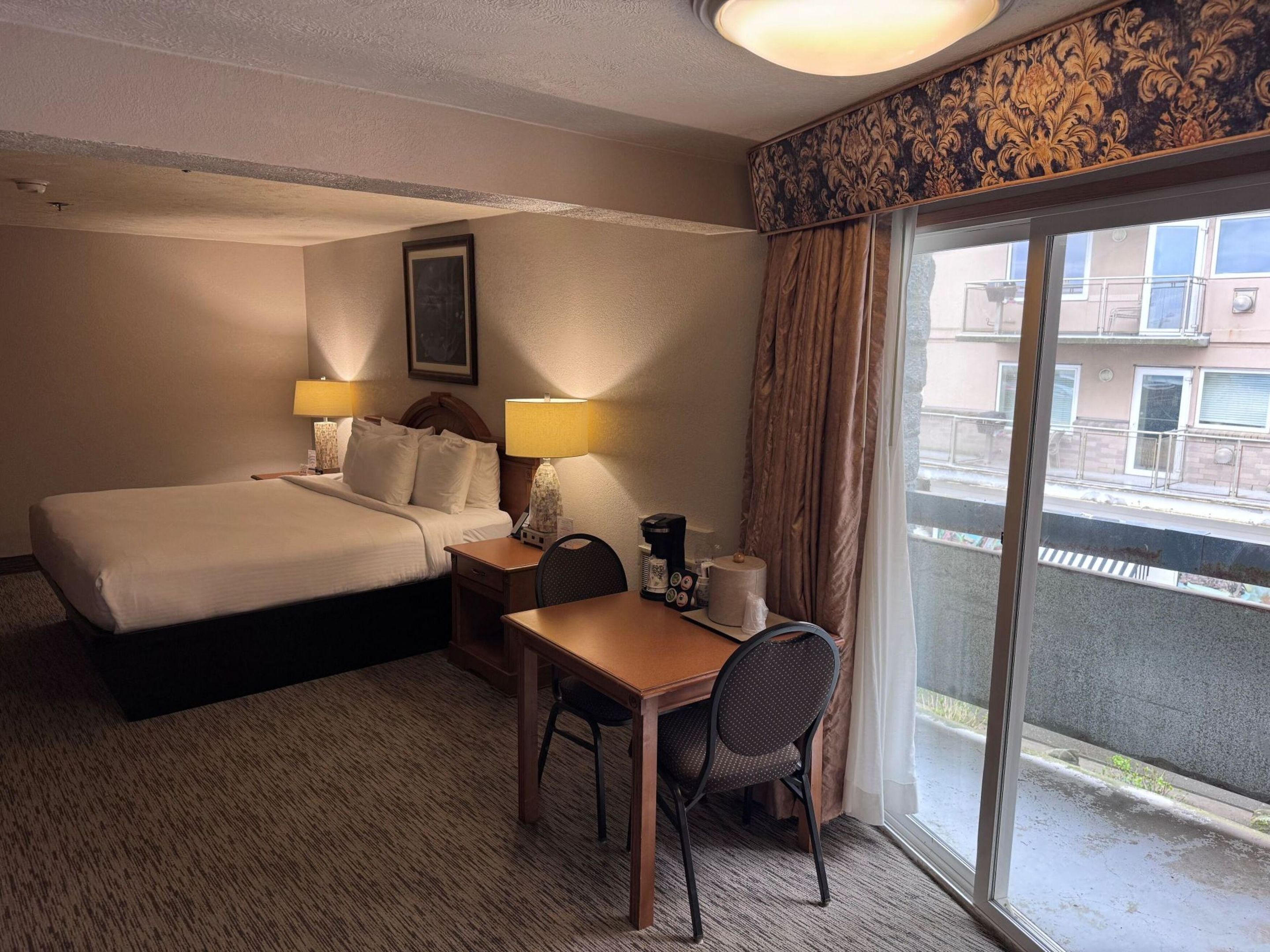 Ein Hotelzimmer mit Bett, Nachttischen, Schreibtisch, Stuhl, Kaffeemaschine und einem Fenster mit Blick auf Gebäude.