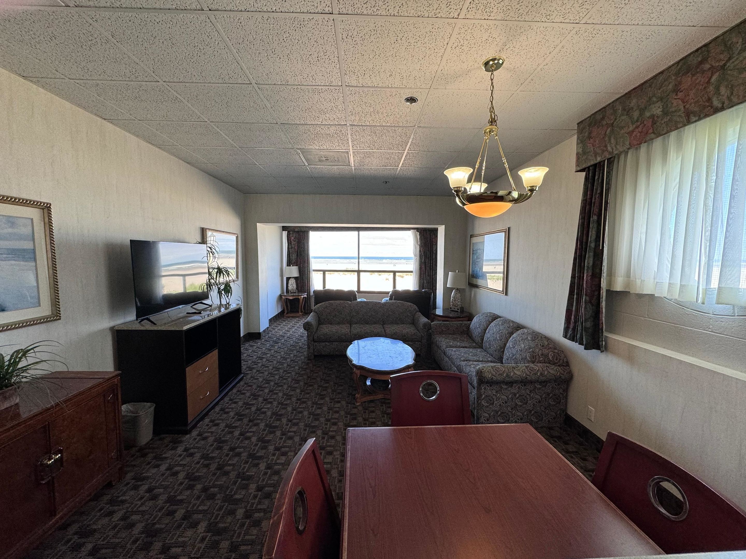 A hotel room with a television, sofa, dining table, and a view of the beach.