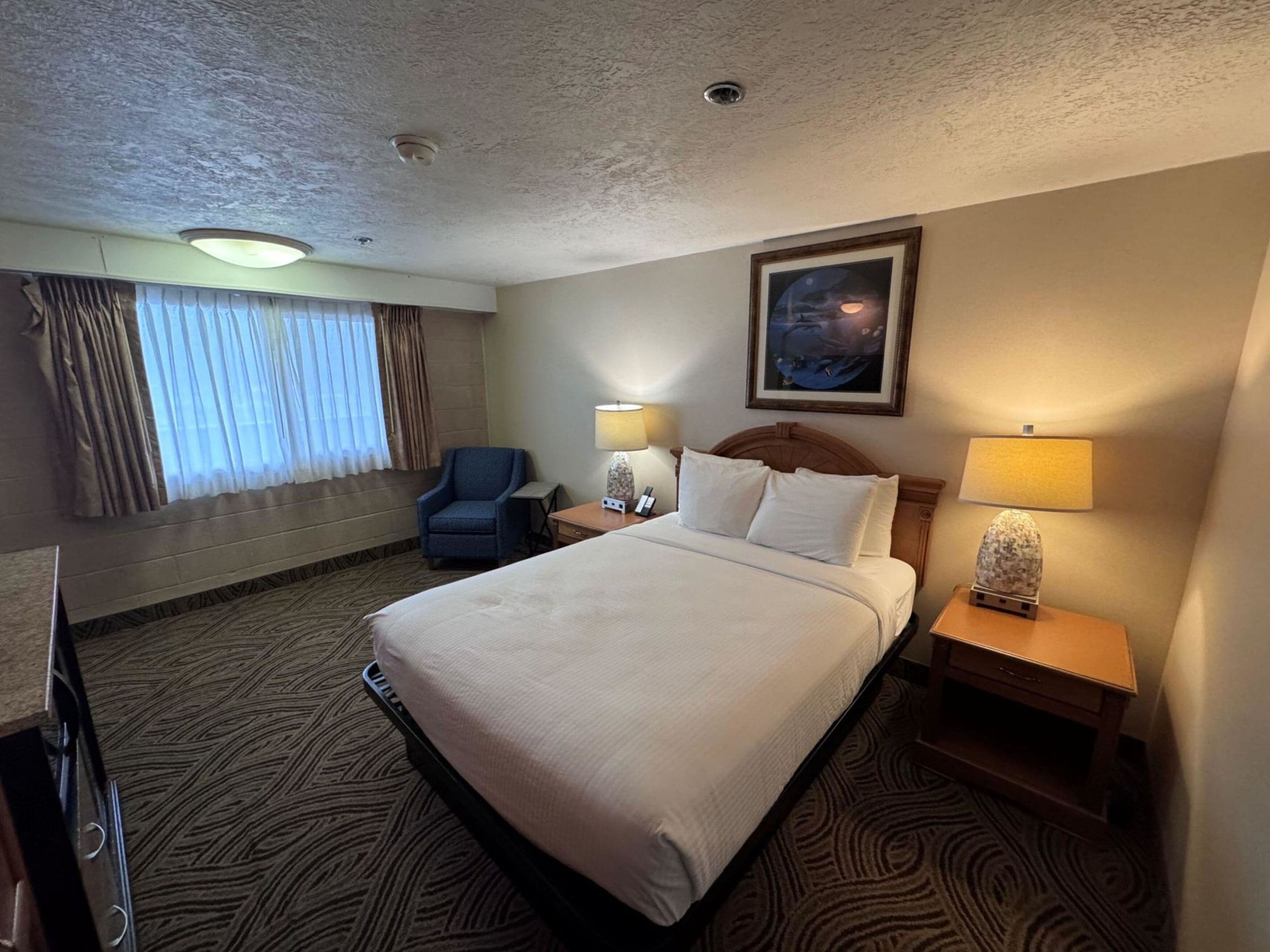 A hotel room with a large bed, two bedside lamps, a chair, a desk, and a framed picture on the wall.