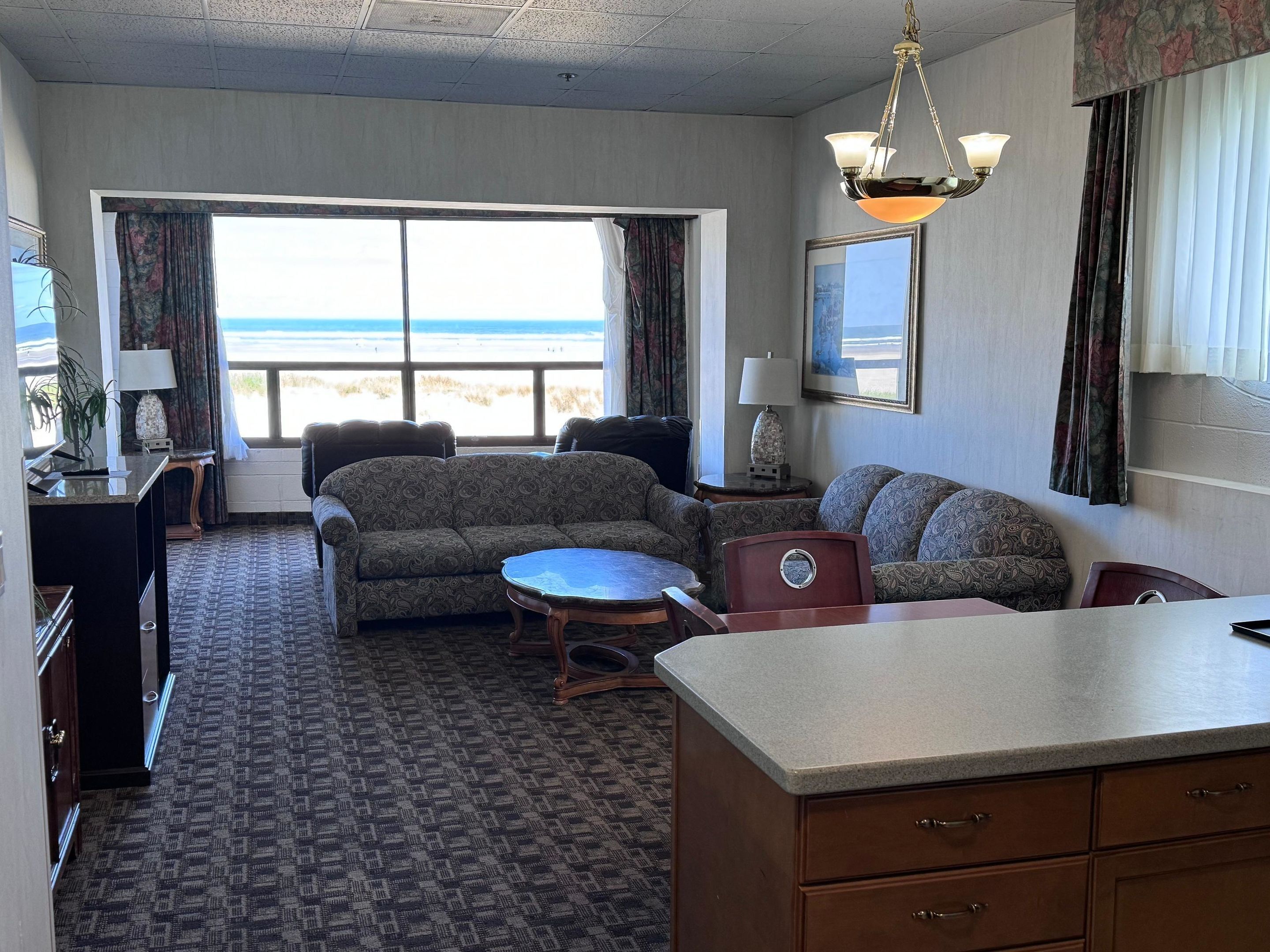 Habitación de hotel con sofá, una mesita, escritorio con lámpara, una silla y vista a la playa.
