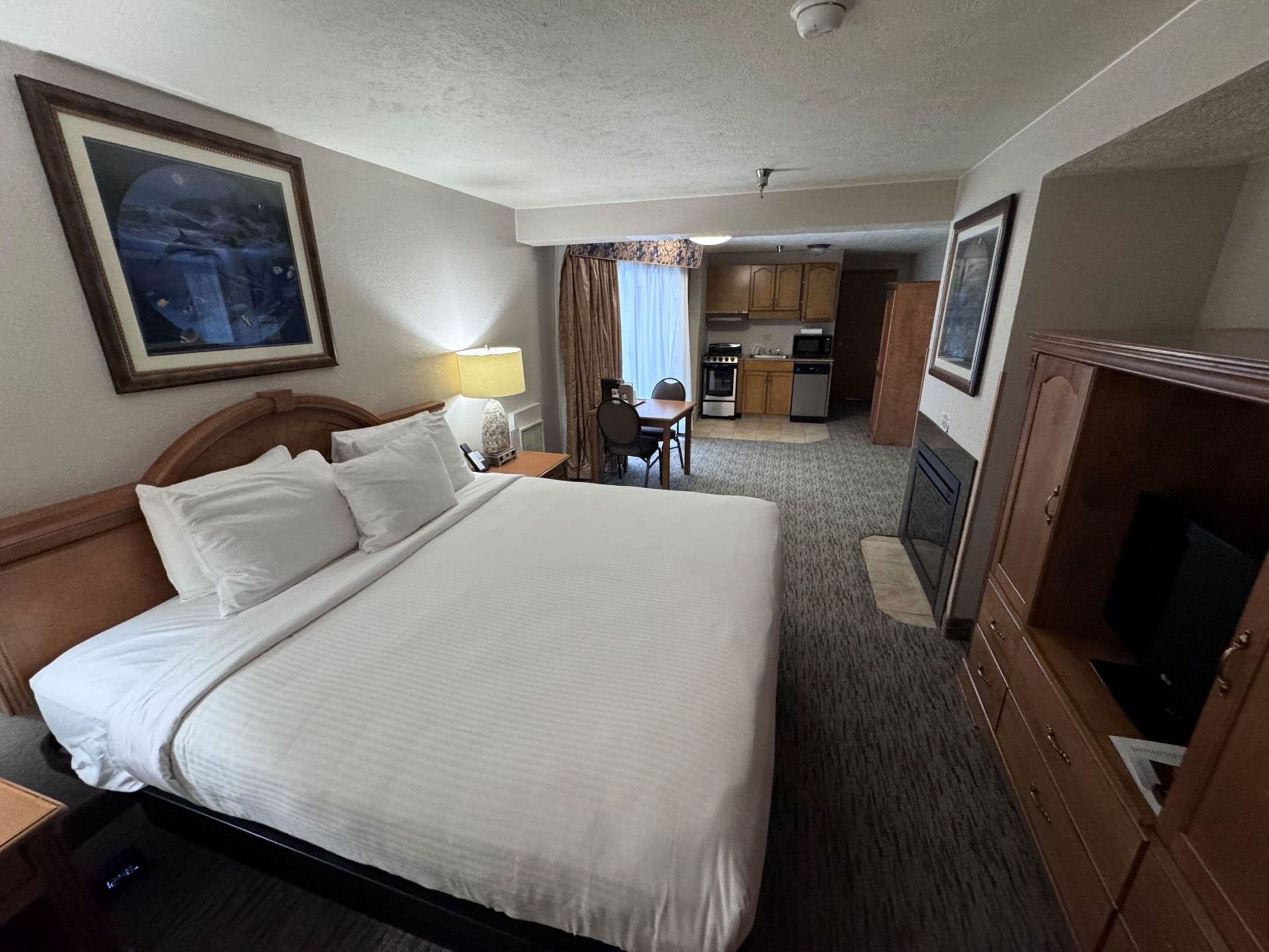 Ein Hotelzimmer mit einem großen Bett, einem Schreibtisch, einem Sessel, einem Fernseher, einer Küchenzeile und einem gerahmten Bild an der Wand.