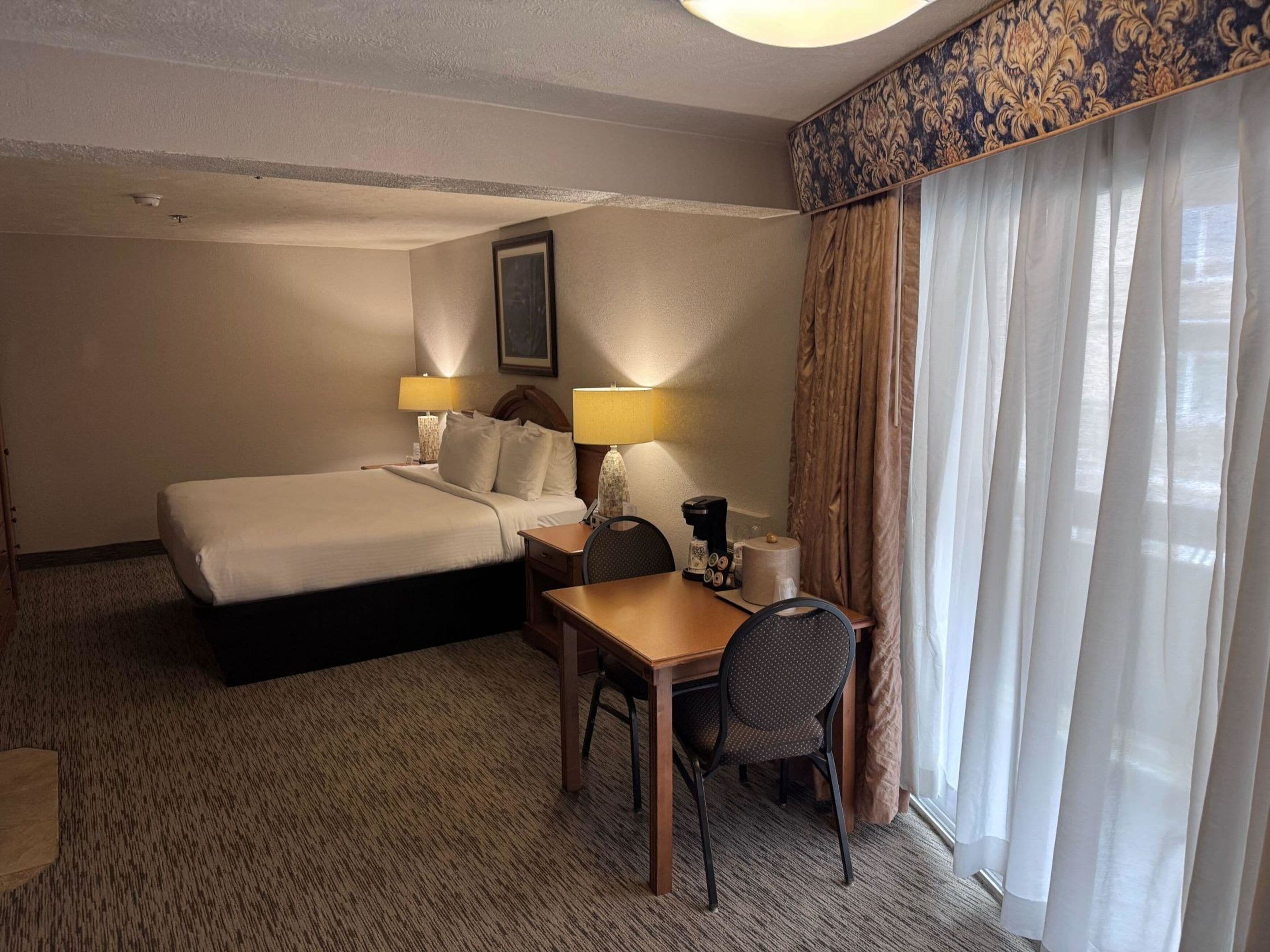 A hotel room with a bed, a desk, a chair, a lamp, a coffee maker, and a window with curtains.