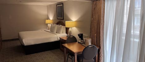 A hotel room with a bed, a desk, a chair, a lamp, a coffee maker, and a window with curtains.