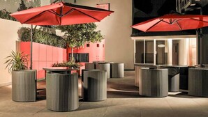 An outdoor seating area with wicker furniture and red umbrellas, surrounded by a low wall and a building entrance. - Asyana Kemayoran Jakarta (Jakarta)