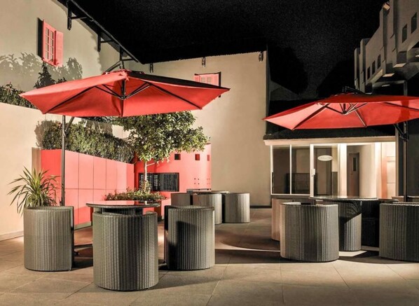 An outdoor seating area with wicker furniture and red umbrellas, surrounded by a low wall and a building entrance. - Asyana Kemayoran Jakarta (Jakarta)