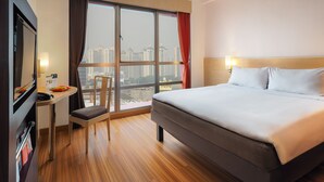 A hotel room with a large bed, a desk with a chair, a TV, and a window with a view of high-rise buildings. - Asyana Kemayoran Jakarta (Jakarta)