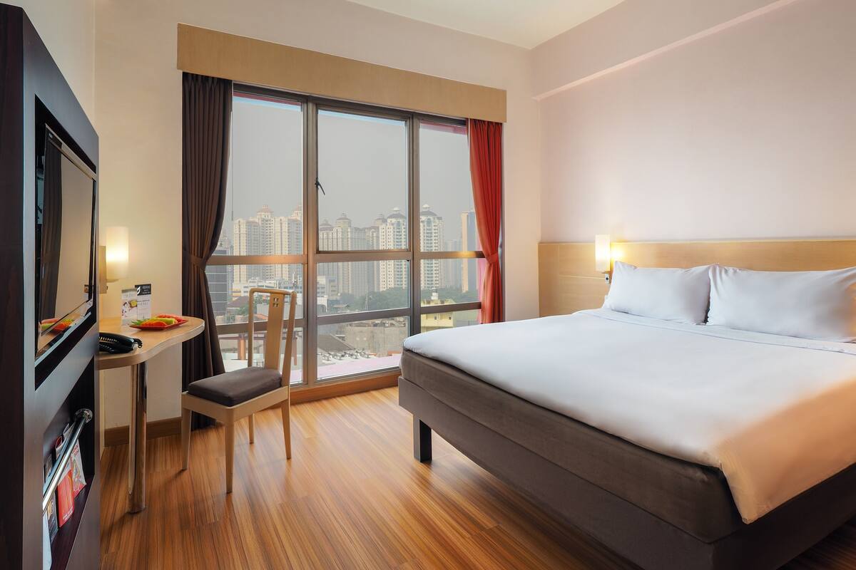 Una habitación de hotel con una cama grande, un escritorio con silla, un televisor y una ventana con vistas a rascacielos.