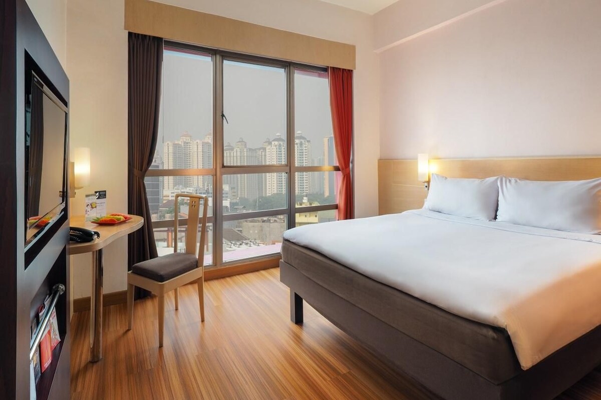 A hotel room with a large bed, a desk with a chair, a television, and a window with curtains.