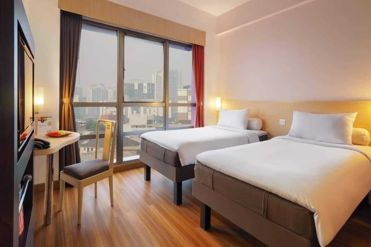 Habitación de hotel con dos camas, un escritorio, una silla y un amplio ventanal con vistas a la ciudad.