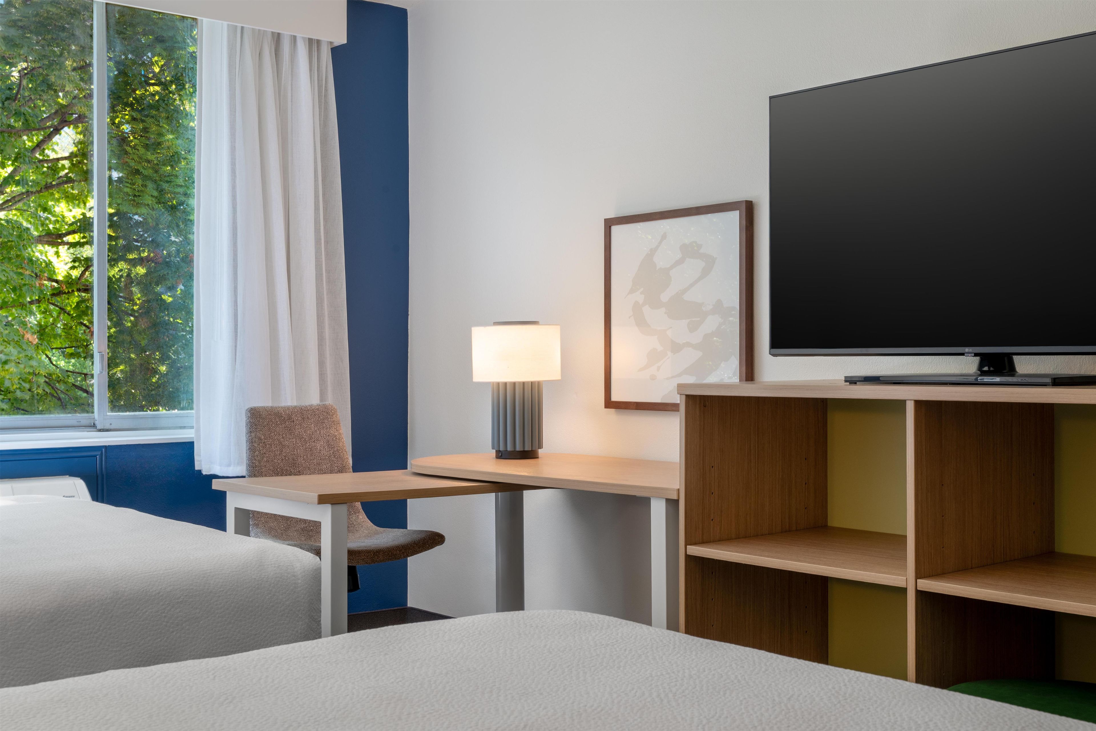 A hotel room with a bed, a desk with a lamp, a television, and a window with curtains.
