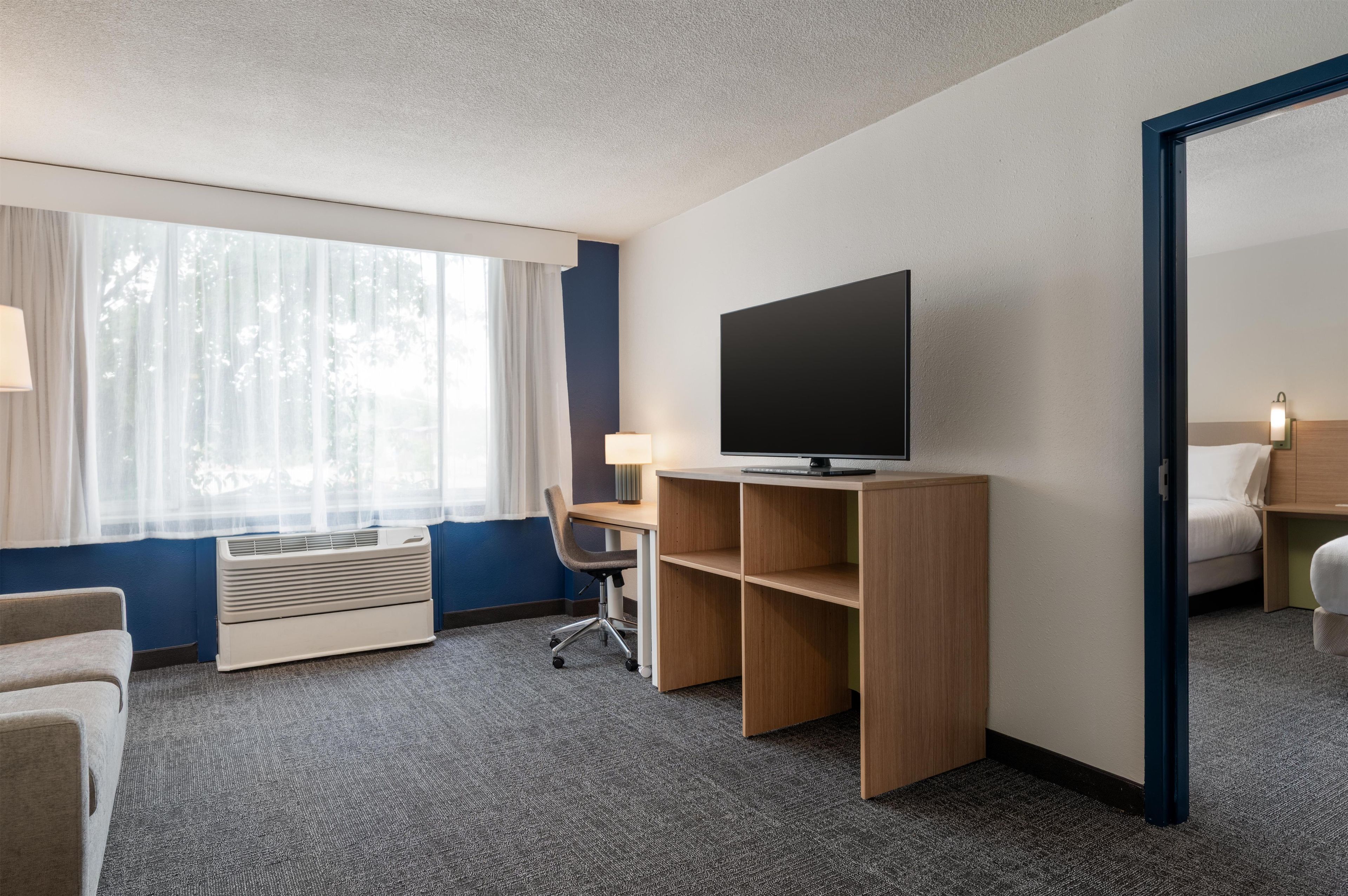 Een hotelkamer met een bed, een bureau met een televisie, een stoel, een raam met gordijnen en een luchtverfrisser aan de muur.
