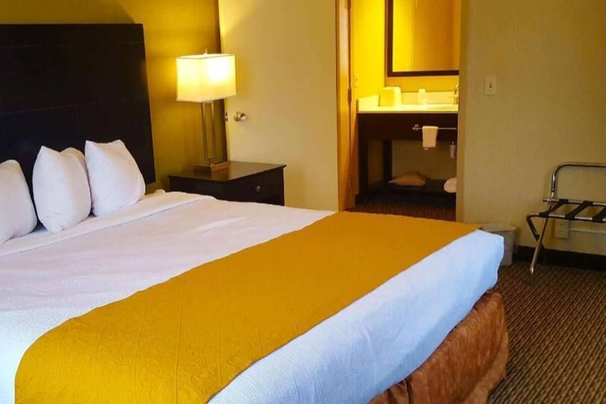 Habitación de hotel con una cama grande, una mesita de noche con lámpara, un baño visible a través de una puerta abierta y una silla.