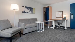 A hotel room with a grey sofa, a small white table, a desk with a chair, a blue door, and a wall-mounted artwork.