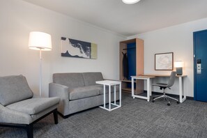 Een hotelkamer met een grijze bank, een witte bijzettafel, een bureau met stoel, een blauwe deur en een kunstwerk aan de muur.