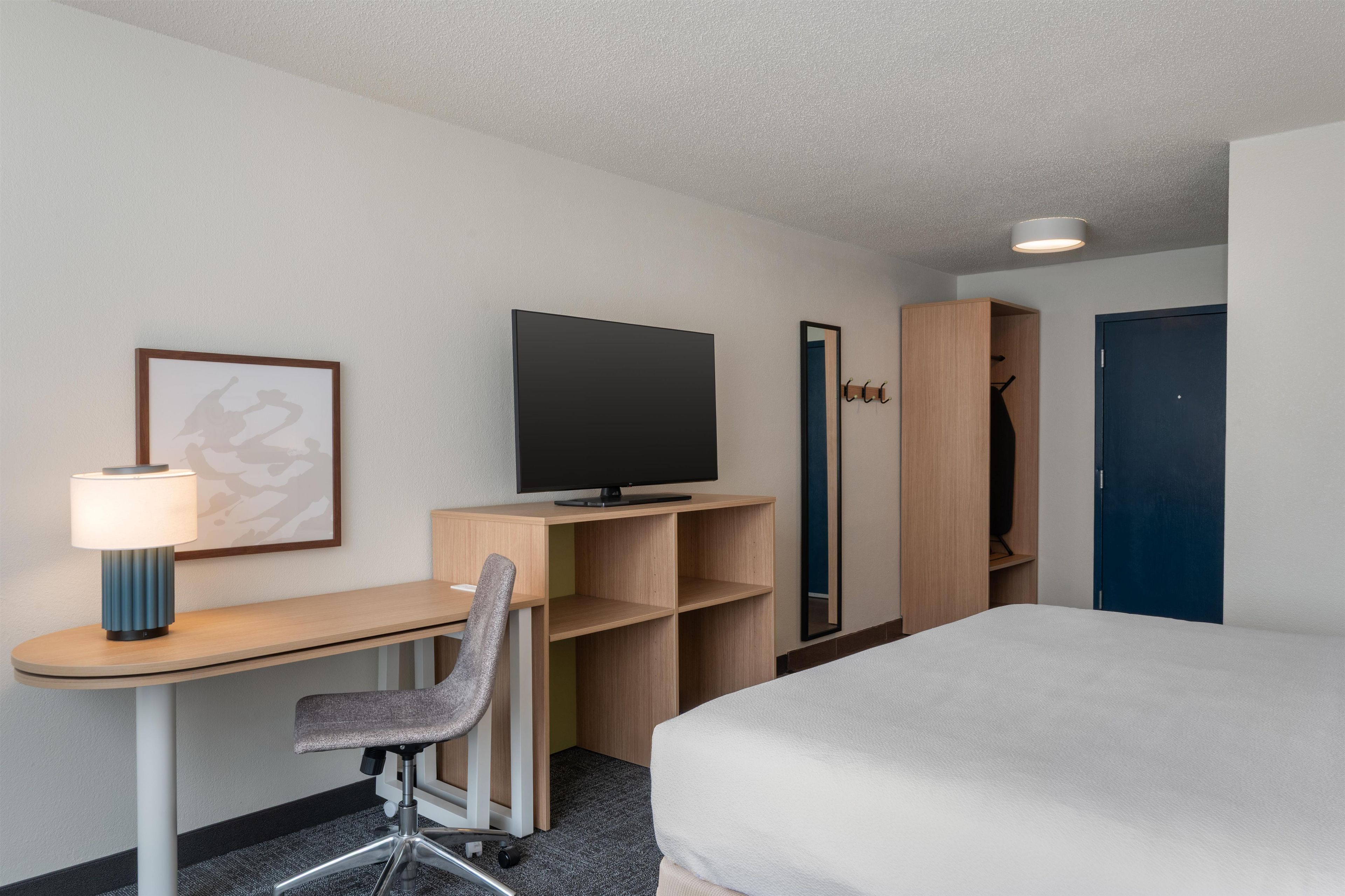 Ett hotellrum med en säng, ett skrivbord med en stol, en tv, en lampa, en klädställning och en garderob.