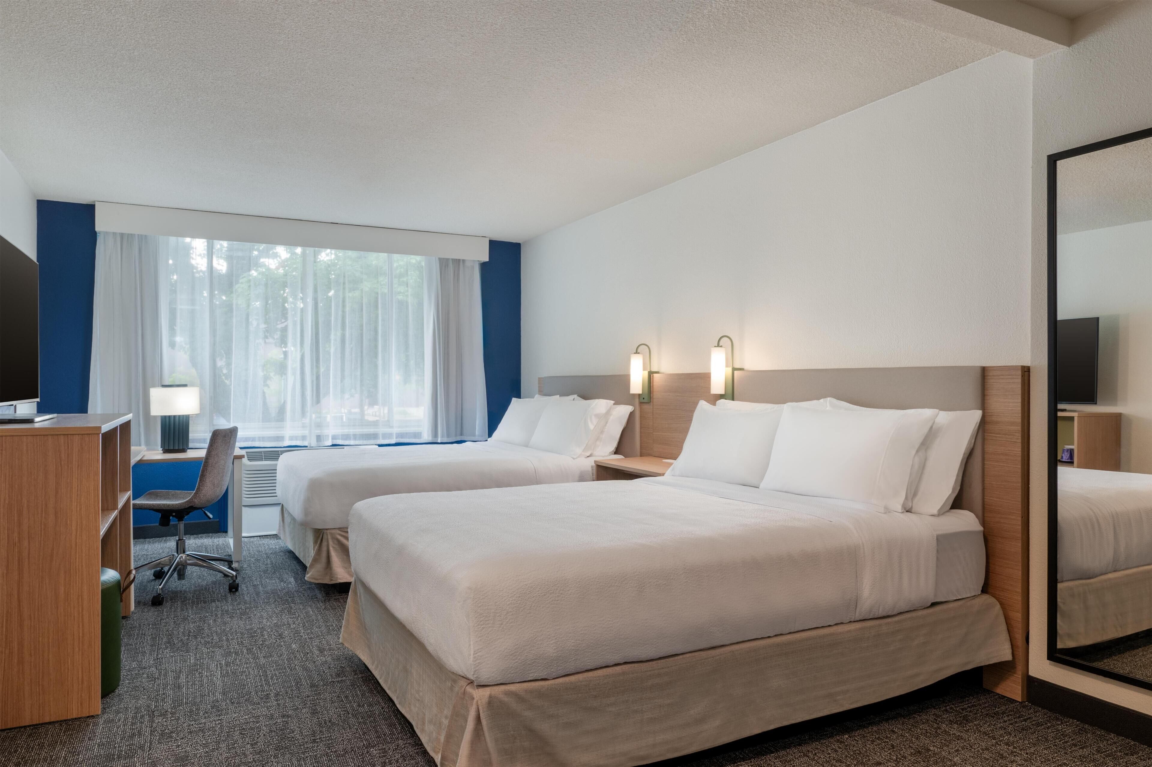 A hotel room with two beds, a desk, a chair, a TV, and a large mirror.