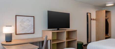 A hotel room with a bed, a desk with a lamp, a television, a coat rack, and a framed picture on the wall.