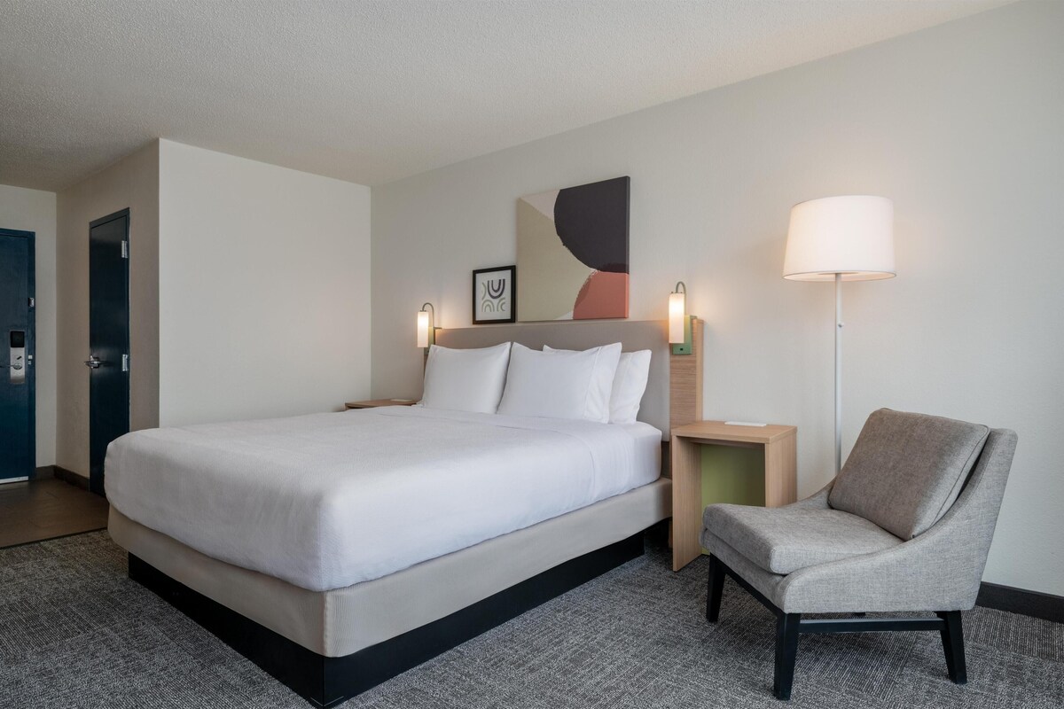 A hotel room with a bed, a chair, a nightstand, a lamp, and a door.