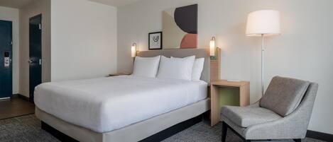 A hotel room with a bed, a chair, a nightstand, a lamp, and a door.