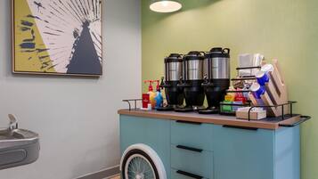 Een kitchenette met een koffiezetapparaat, een waterkoker en een waterdispenser. Er is een spoelbak en een rolstoeltoegankelijke kruiwagen.