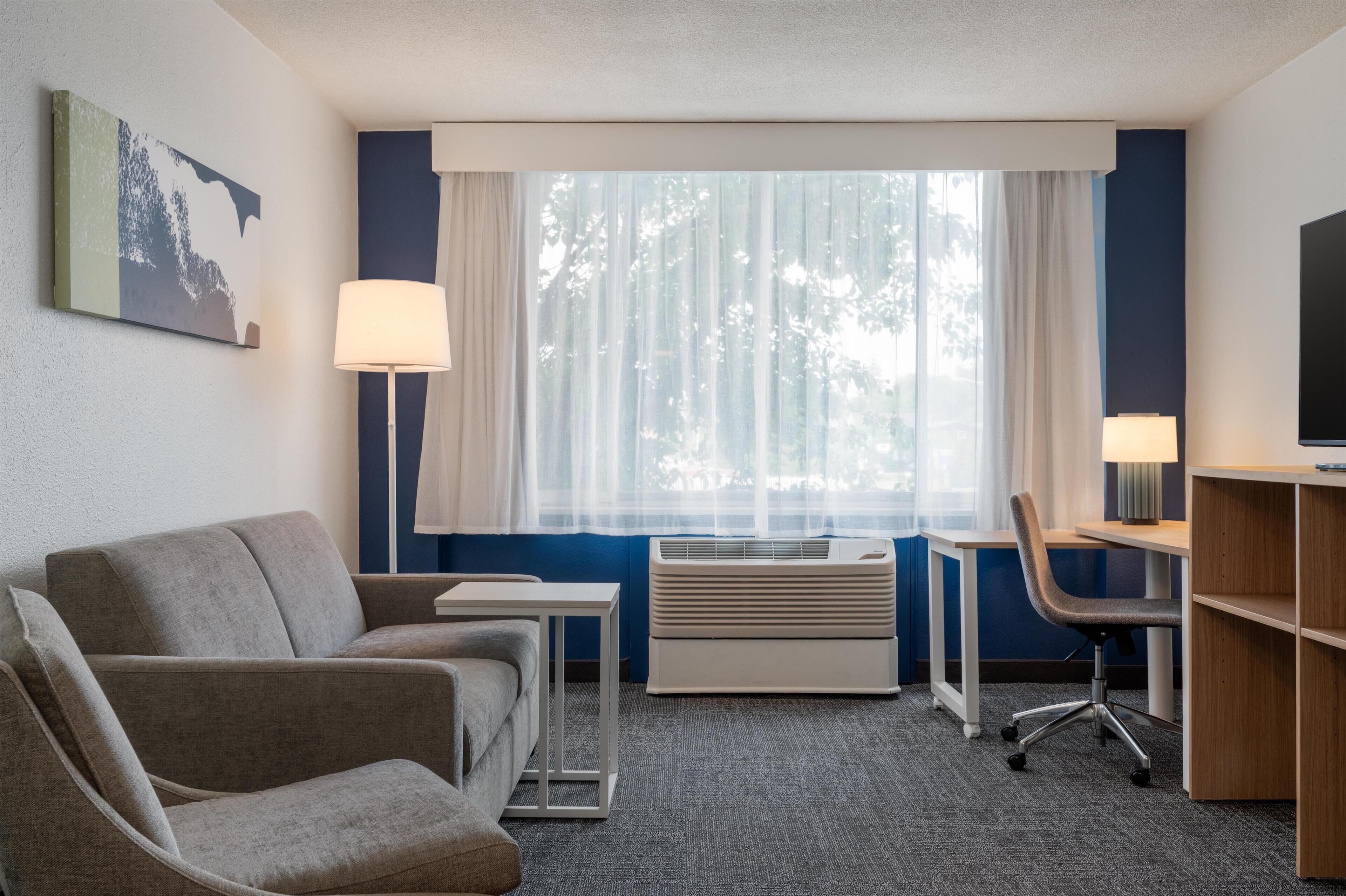 A hotel room with a sofa, a desk, a chair, a TV, and a window with curtains.