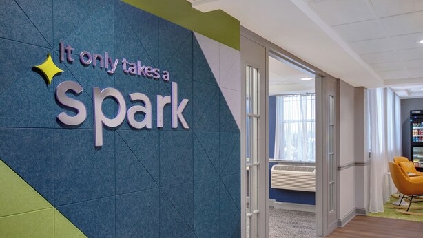 A sign with the text 'It only takes a spark' and a yellow star, a glass door leading to an interior space, and a yellow chair. - Spark by Hilton Watertown (Watertown)