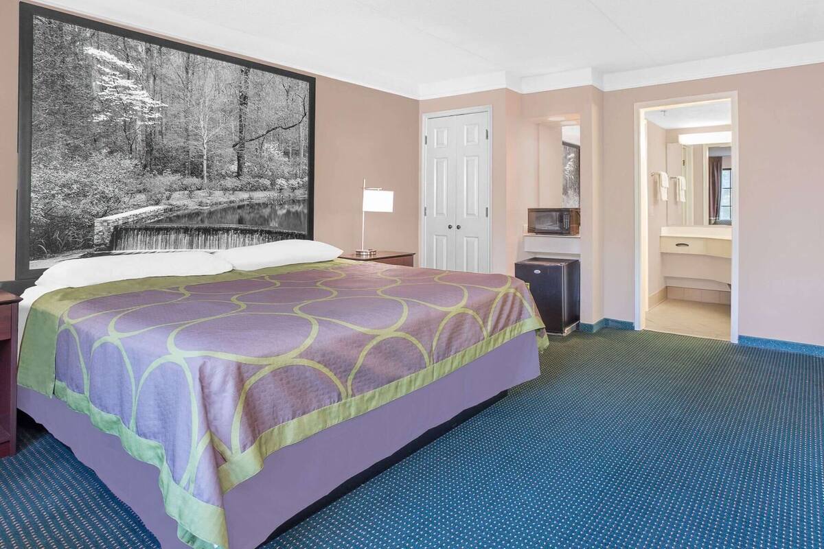 A hotel room with a bed, a large framed picture, a door, a microwave, and a bathroom visible through an open door.