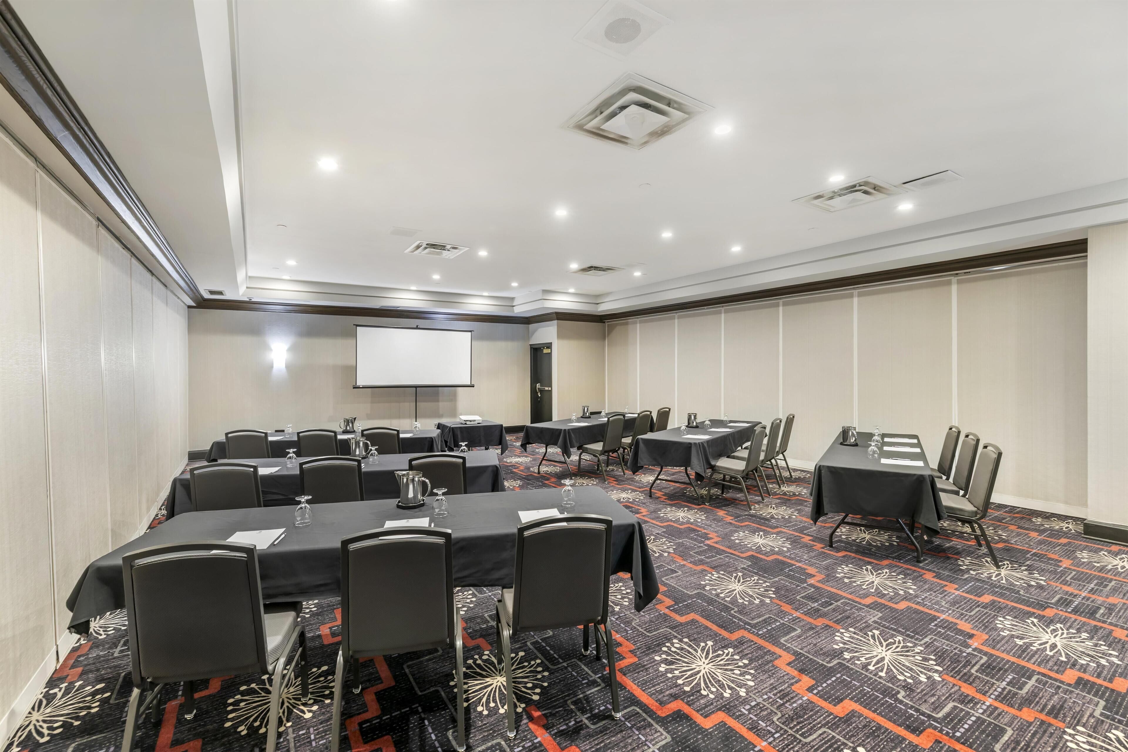 A conference room with tables and chairs, a projector screen, and a carpeted floor.