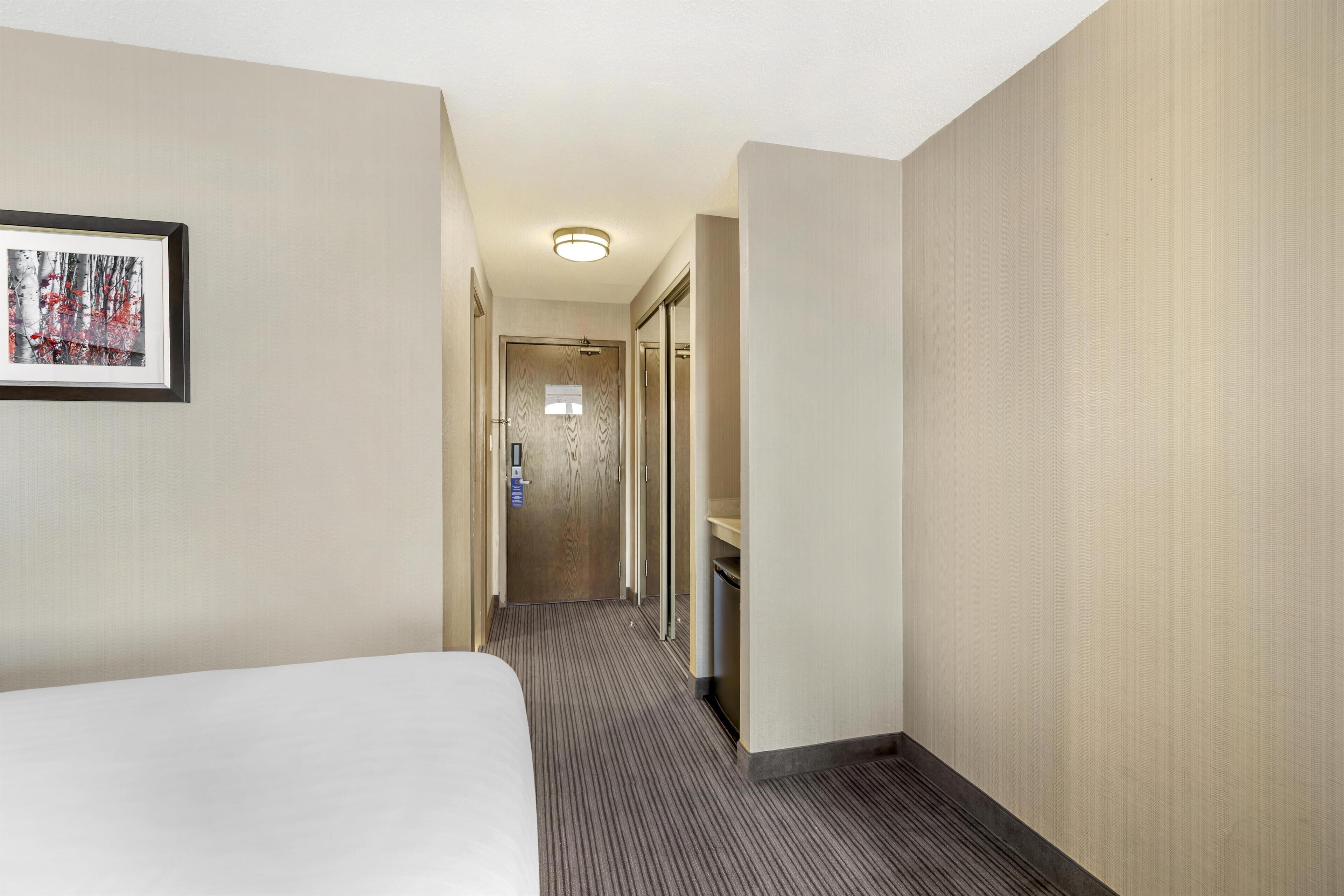 Ein Hotelgang mit einem Bett auf der linken Seite, einem Badezimmer mit Spiegel und Waschbecken auf der rechten Seite und einer Tür am Ende.