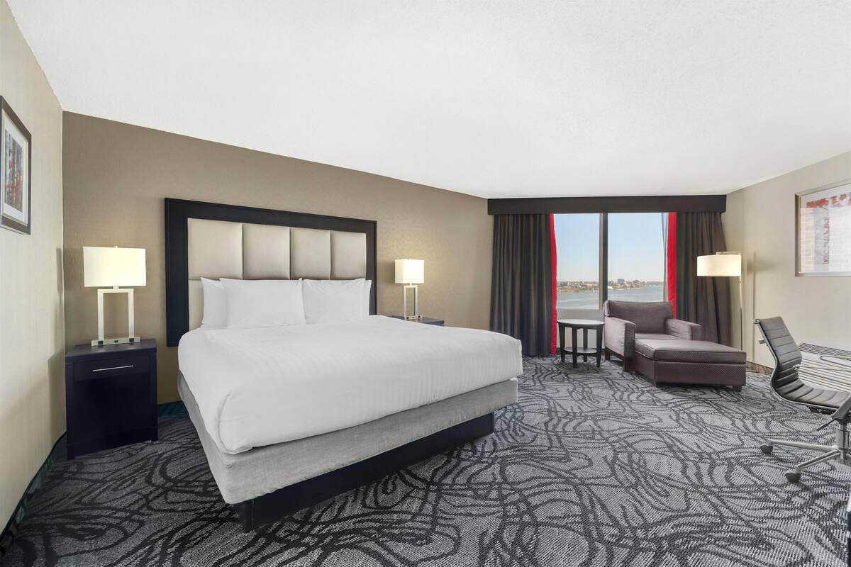 Una habitación de hotel con una cama grande, dos mesitas de noche con lámparas, una pequeña mesa con una silla y vistas a la ciudad a través de grandes ventanales.