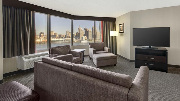 A hotel room with a large window offering a city skyline view, a flat-screen TV, a sofa, two armchairs, a small table, and a lamp. - Best Western Plus Waterfront Hotel (Windsor)