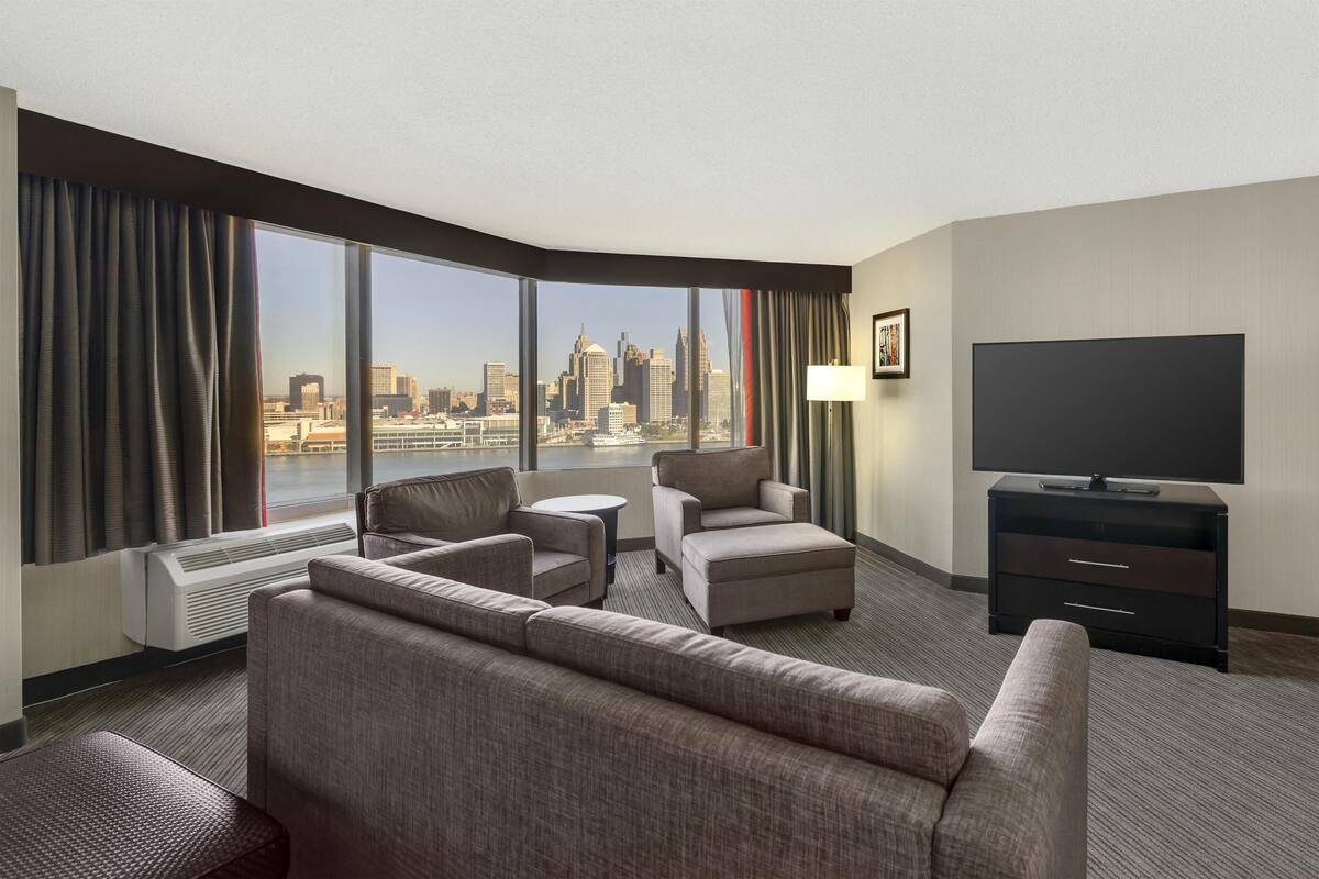 Habitación de hotel con un ventanal grande que ofrece vistas a un horizonte urbano, un televisor de pantalla plana, un sofá, dos sillones, una mesita y una lámpara.