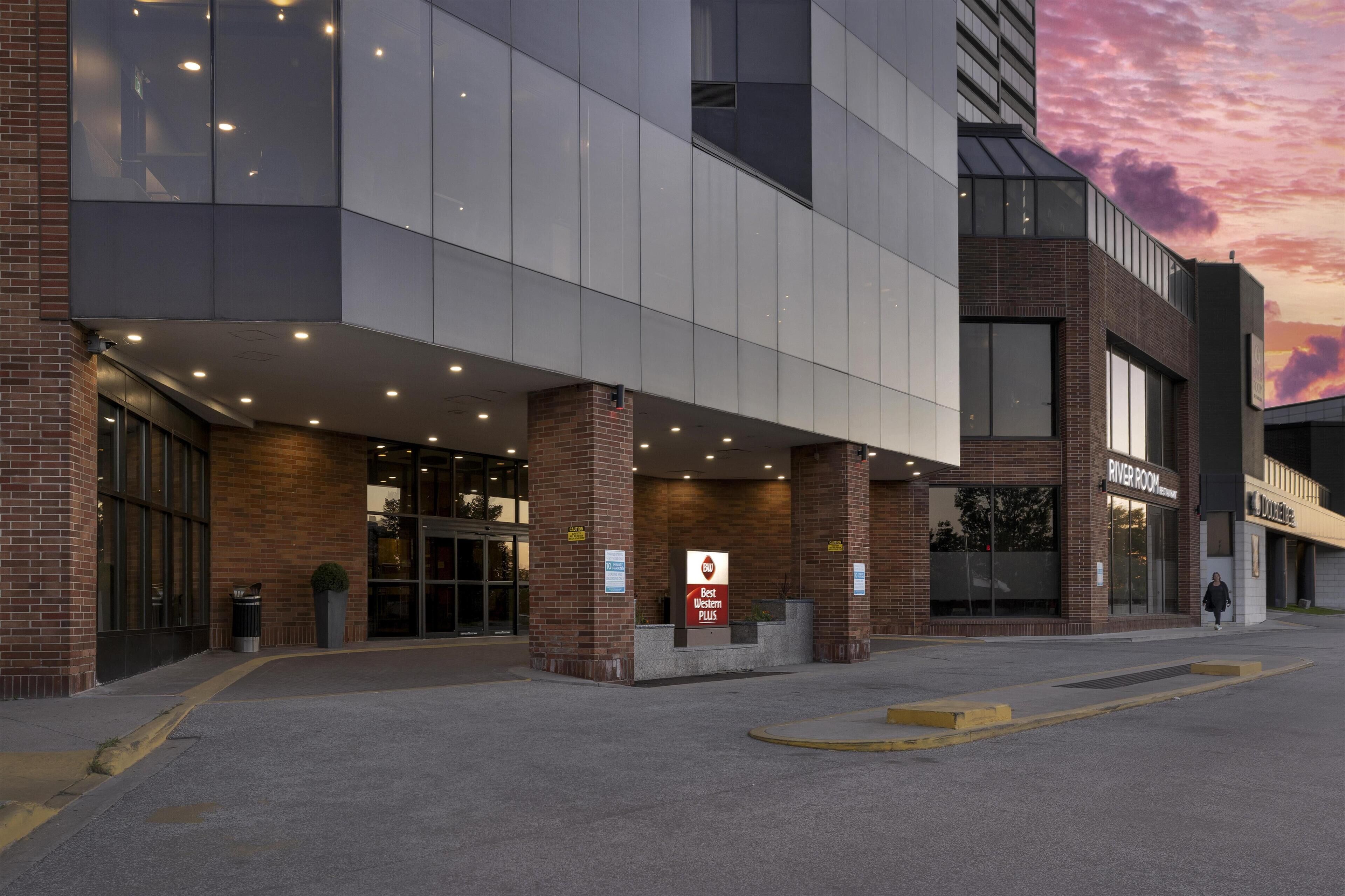 A modern hotel entrance with a glass facade, a brick lower level, and a sign for the River Room.