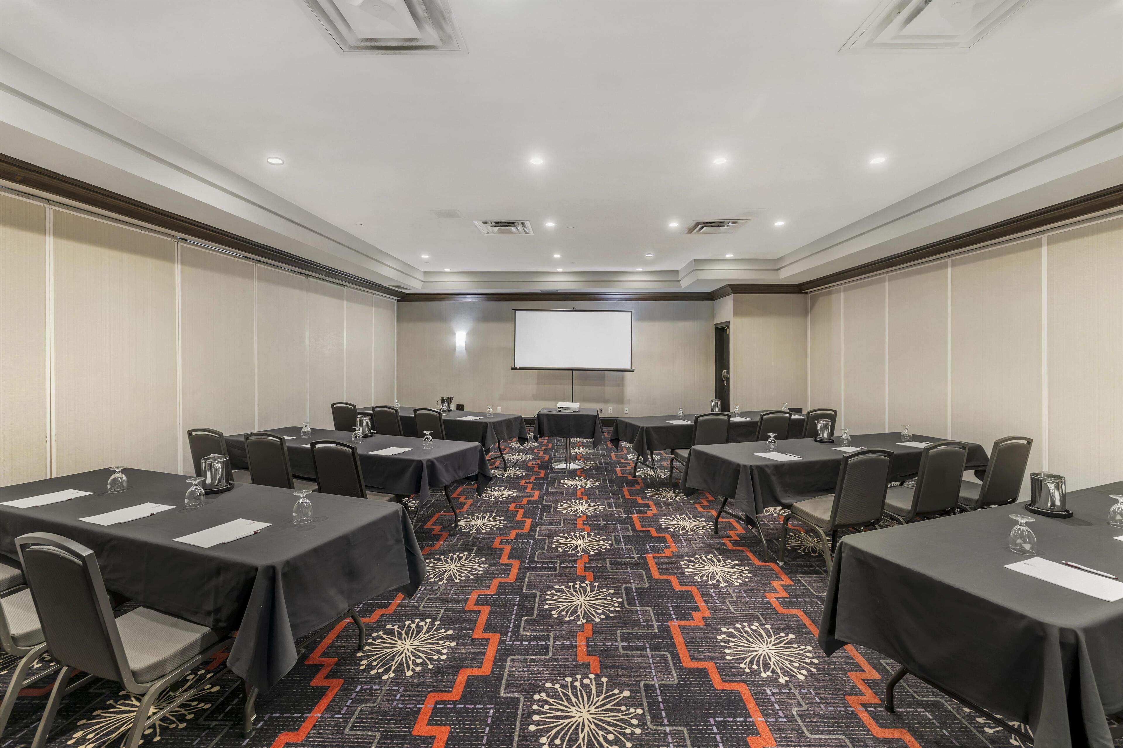 A conference room with tables, chairs, a projector screen, and a patterned carpet.