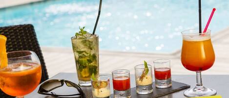 A table with various drinks and glasses, a pair of sunglasses, and a yellow napkin, set against a poolside view.